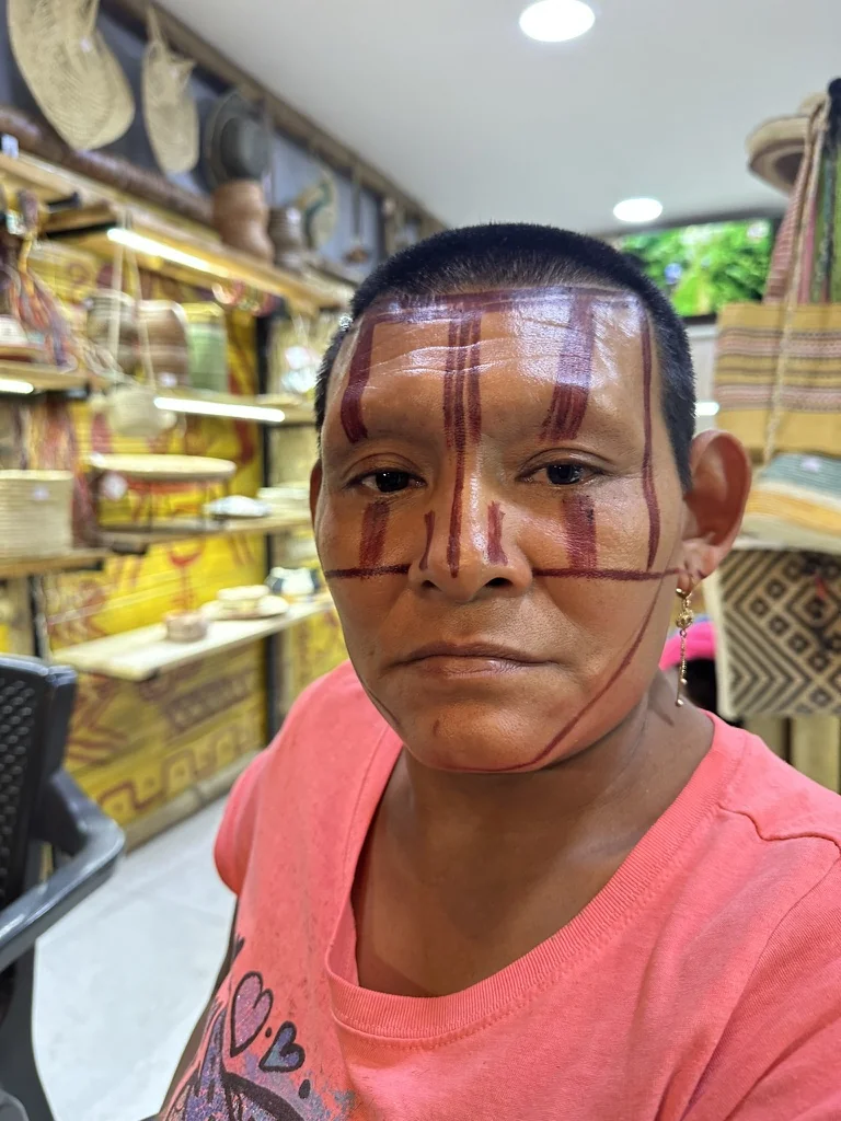 Indigenous ambassador with traditional red face markings at Dulce Amazónica store, artisan goods on shelves behind