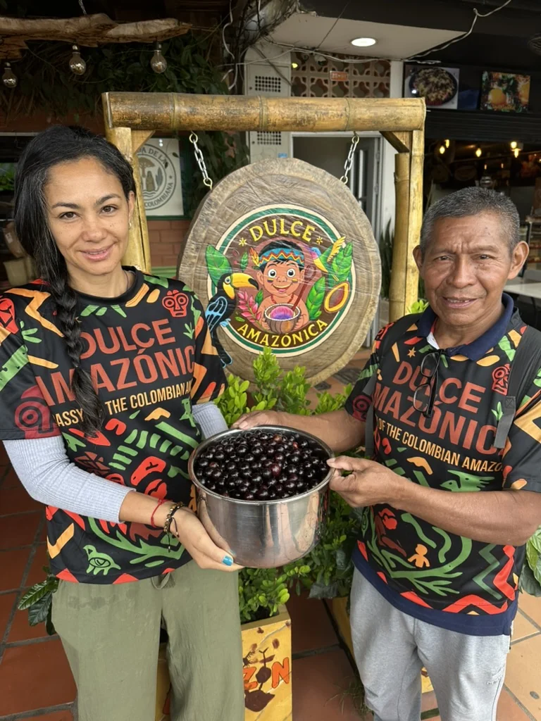 Johana and indigenous ambassador holding a bowl of Jabuticaba fruit in front of Dulce Amazónica sign, Guatapé